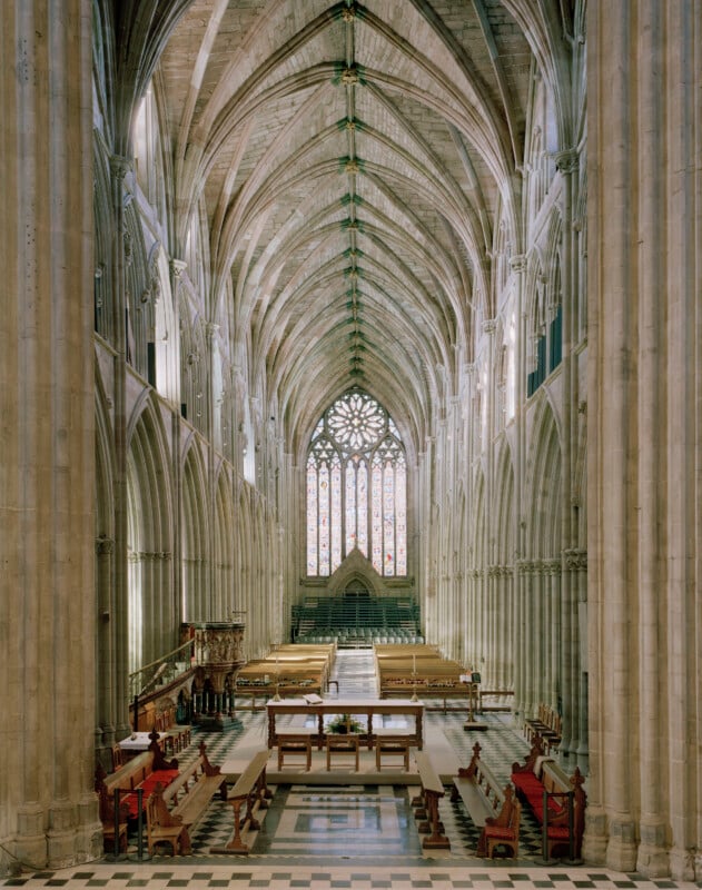 The interior of a Gothic cathedral features a long nave with vaulted ceilings, stone columns, wooden pews, and a stained-glass window at the far end.