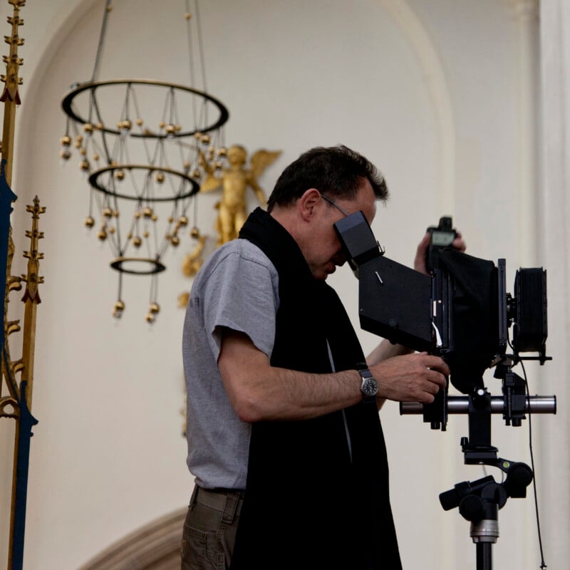 A man in a grey t-shirt and black scarf looks through the viewfinder of a large-format camera mounted on a tripod inside a building.