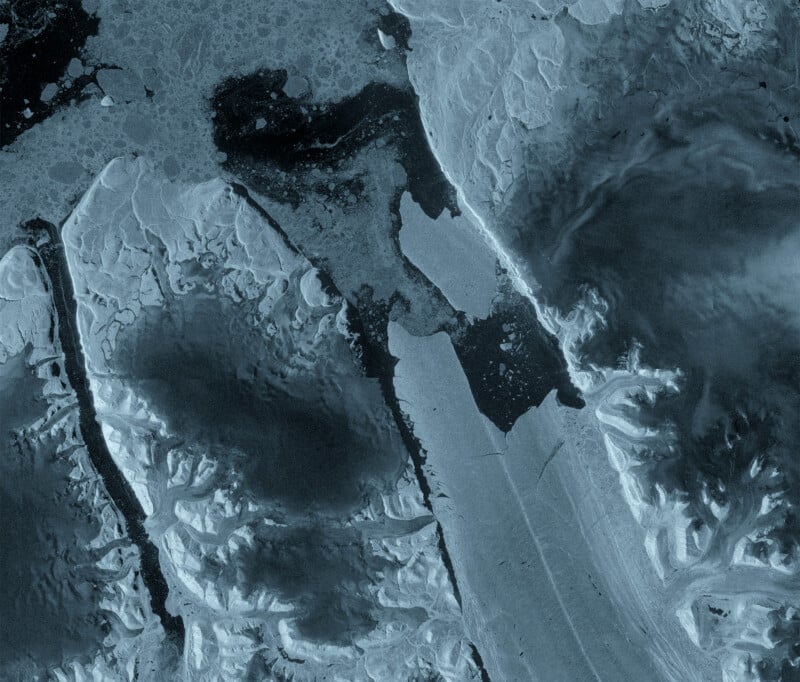 A satellite view of a glacier flowing into dark water, surrounded by rugged, snow-covered terrain and scattered sea ice.
