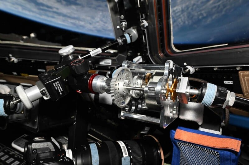A mechanical gear assembly mounted inside a spacecraft window, with a camera lens visible below and the Earth's horizon in the background.
