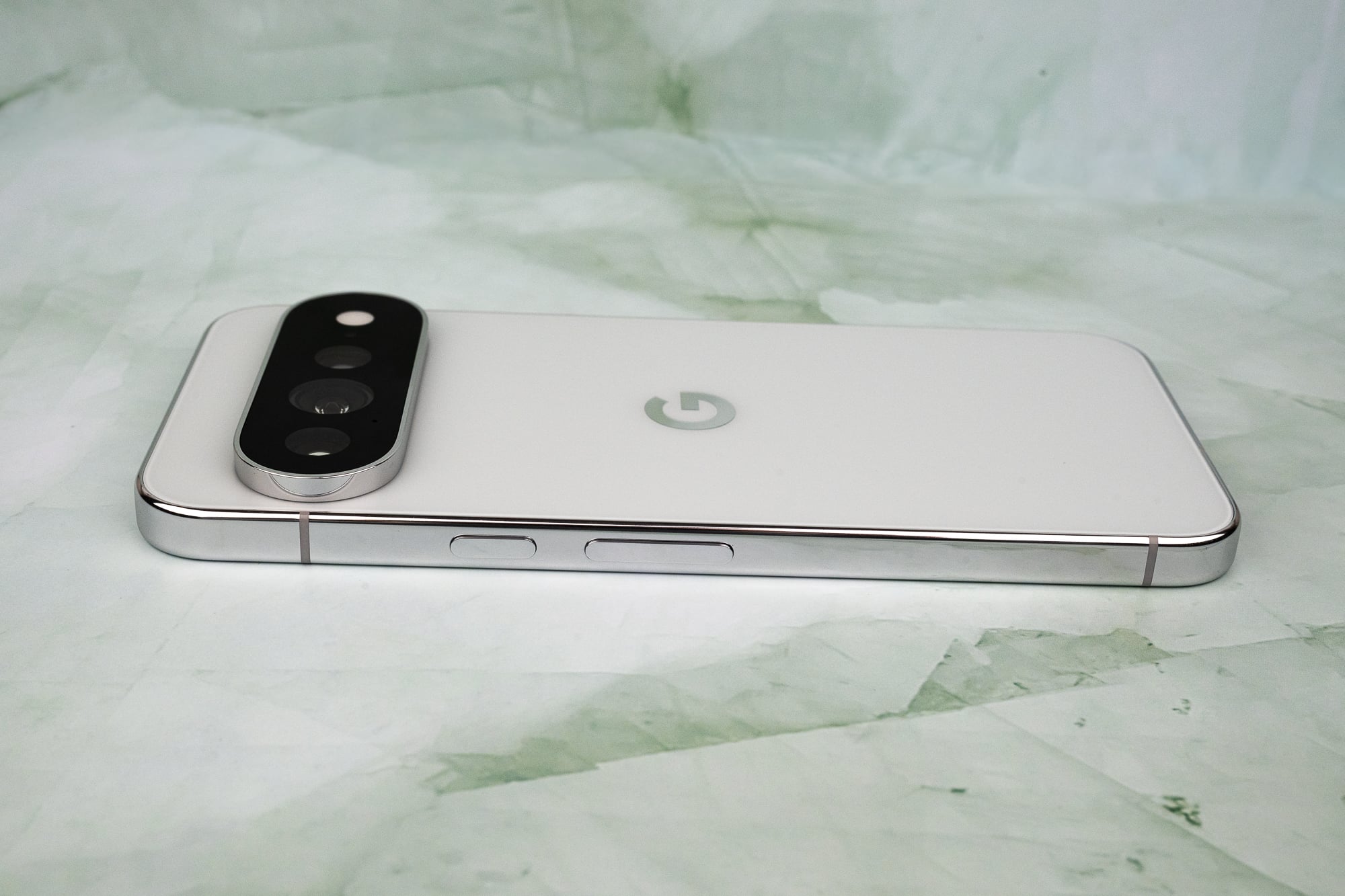 A white smartphone with a prominent, pill-shaped camera module on the back lies on a light green, marbled surface.