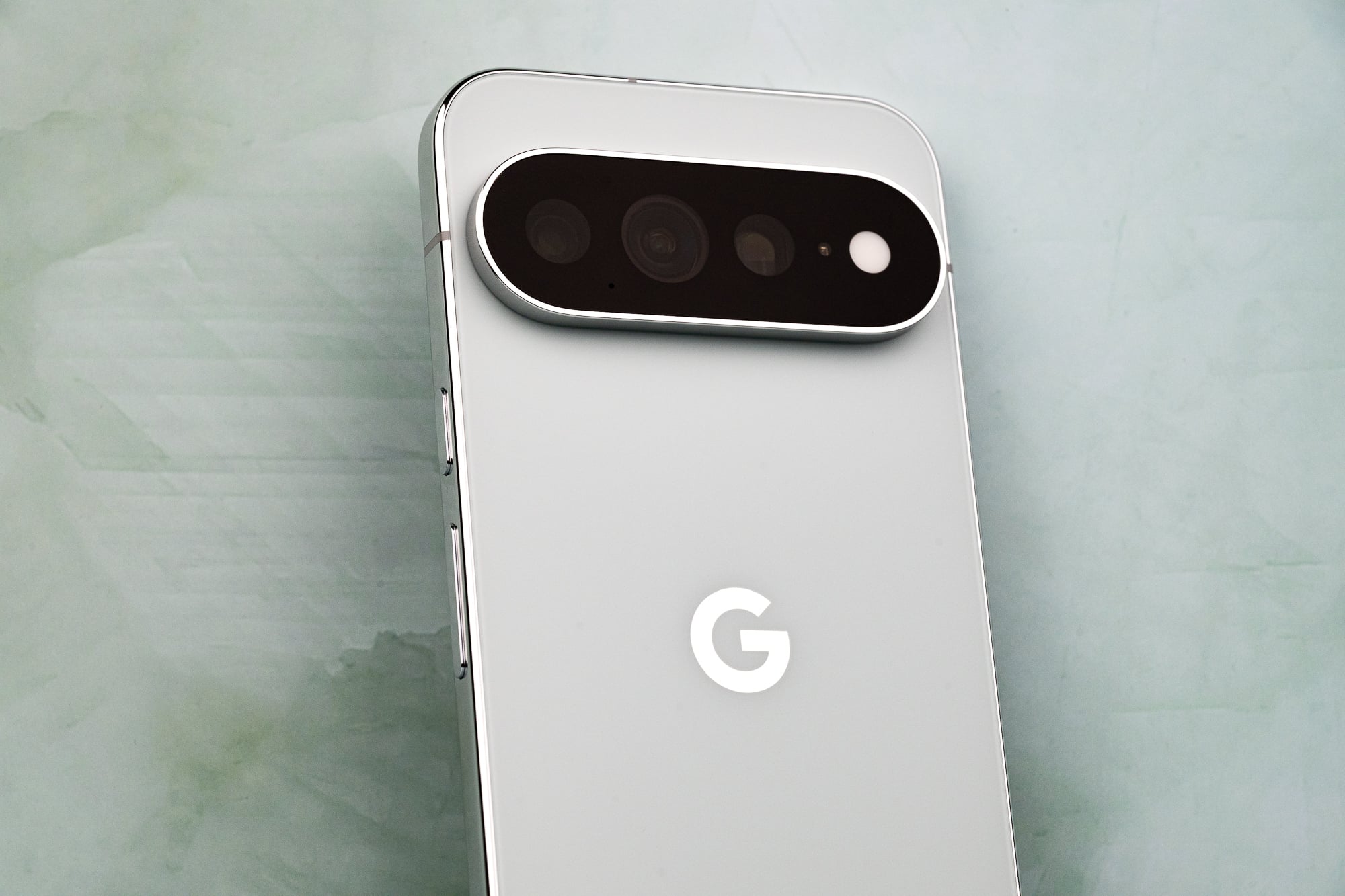 A close-up, angled view of the back of a light gray smartphone featuring a prominent, horizontal, pill-shaped camera module with three lenses and a flash, and a white "G" logo centered on the body.