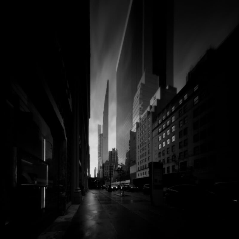 A low-angle, black-and-white view of a city street lined with tall skyscrapers and buildings, with a narrow sky visible between them.