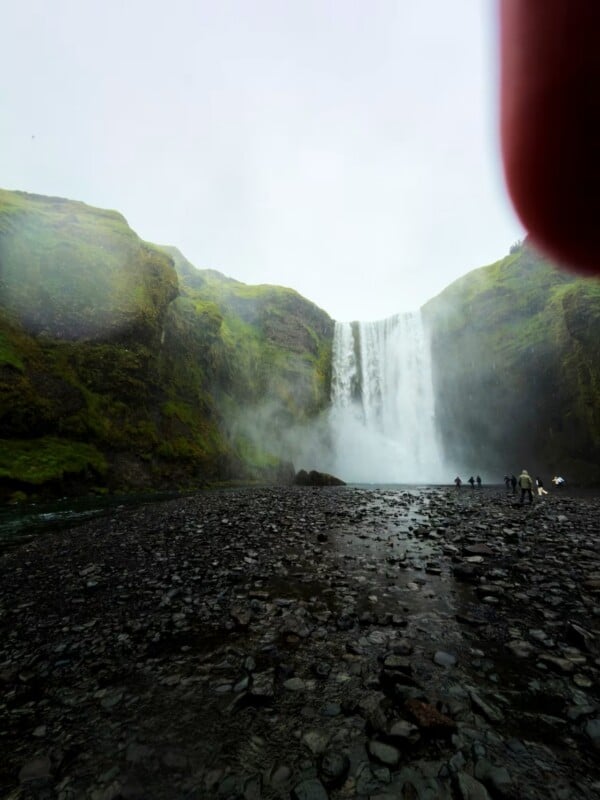 A large waterfall cascades down a green, mossy cliff face onto a dark, rocky riverbed with several people standing nearby.