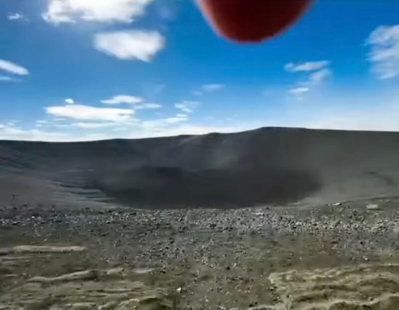 A wide, bowl-shaped volcanic crater with dark, rocky slopes under a bright blue sky with scattered clouds.