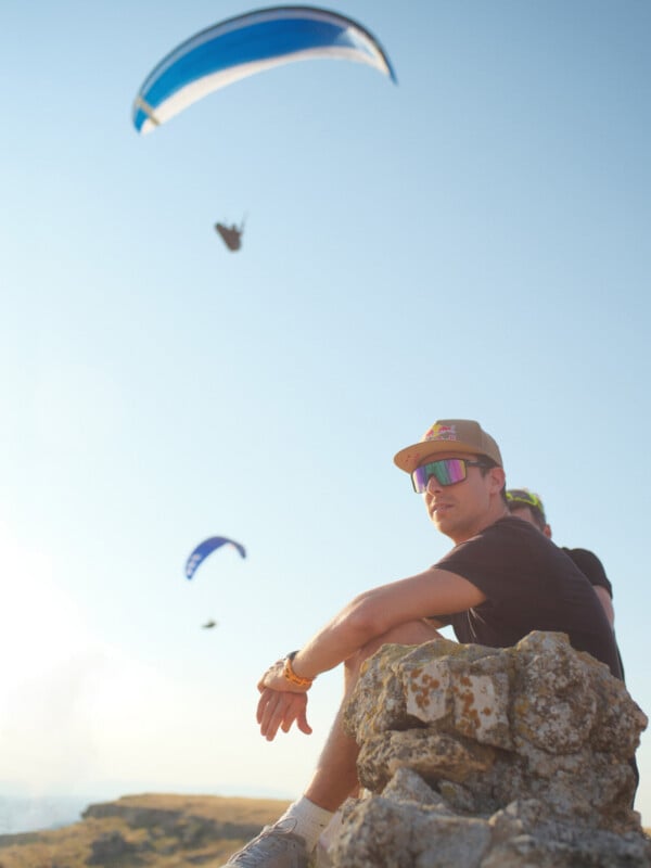 A man wearing a baseball cap and sunglasses sits on a rocky ledge, looking out as paragliders fly in the sky above him.
