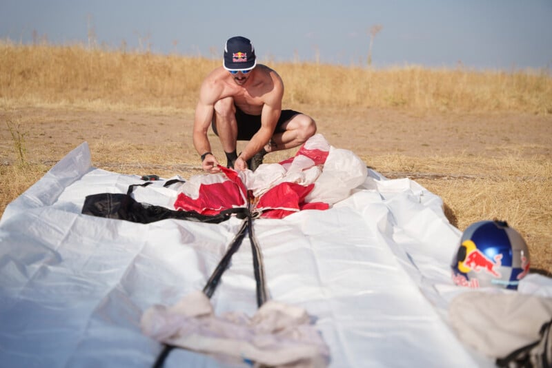 A shirtless man wearing a Red Bull cap and sunglasses kneels on the ground, carefully packing a red and white parachute on a white mat, with a matching helmet nearby.