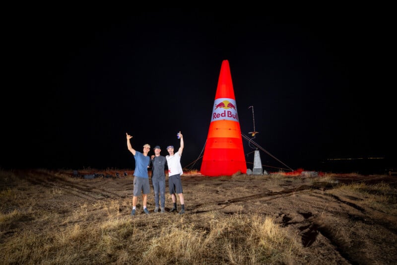 Three men stand in a field at night in front of a large, illuminated, orange Red Bull inflatable cone.