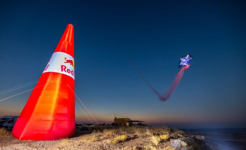 A person in a wingsuit with a red smoke trail flies near a large, inflatable red Red Bull pylon at dusk.