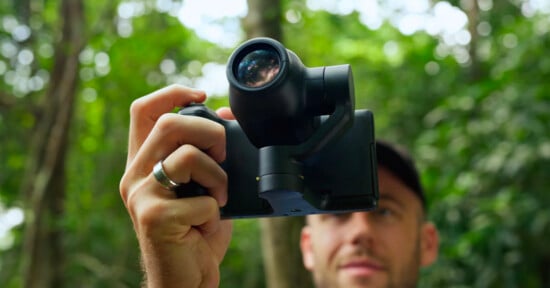 A person holds a black gimbal-mounted camera in front of their face in a wooded area.