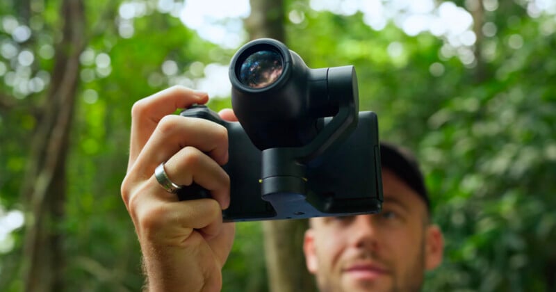 A person holds a black gimbal-mounted camera in front of their face in a wooded area.