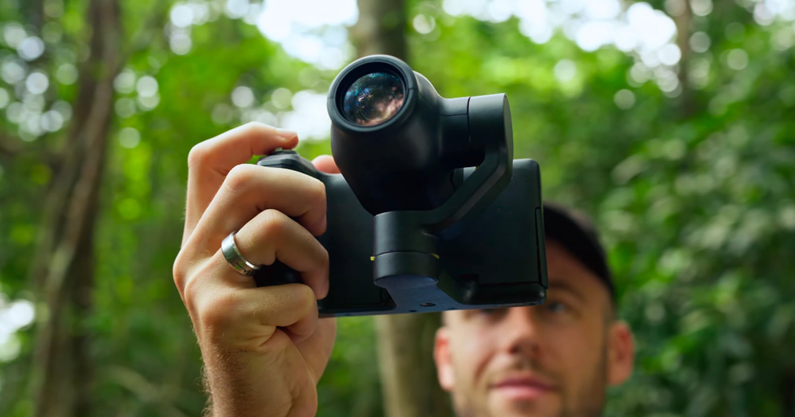 A person holds a black gimbal-mounted camera in front of their face in a wooded area.
