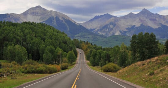 A paved road stretches into the distance toward a range of large, rugged mountains under a cloudy sky, flanked by dense green forests.