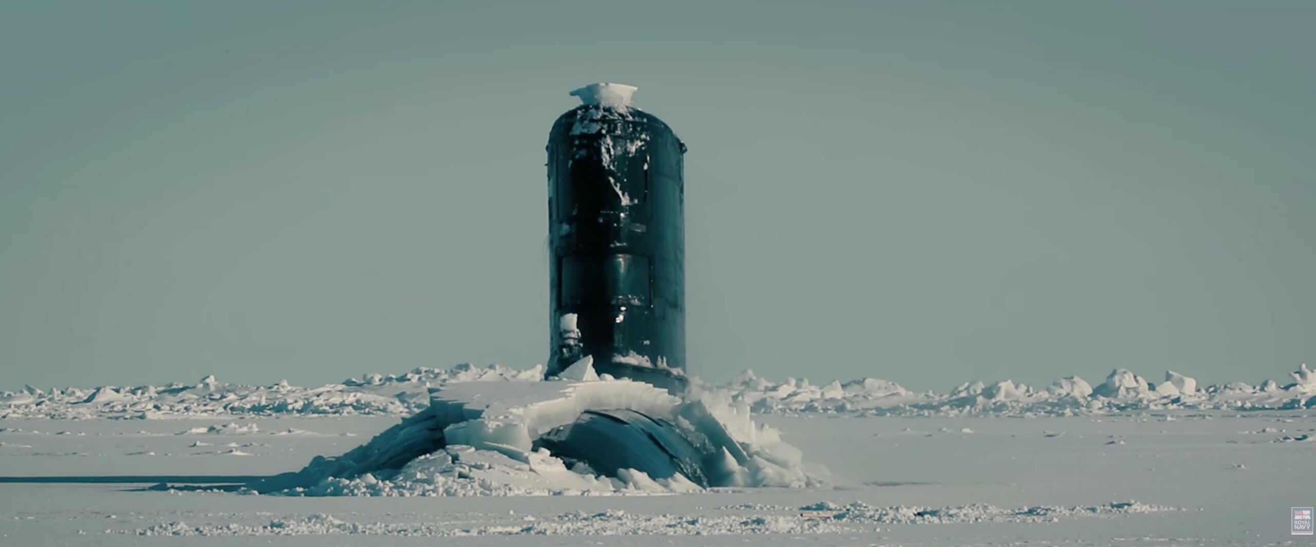 A submarine conning tower breaks through a thick layer of ice in a frozen, snow-covered landscape.