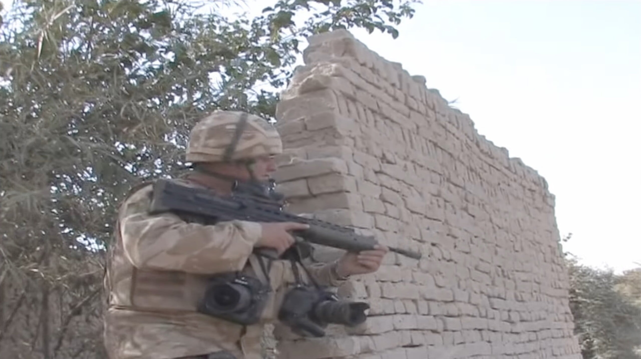 A soldier in desert camouflage uniform stands by a brick wall, holding a rifle and carrying two cameras around their neck.