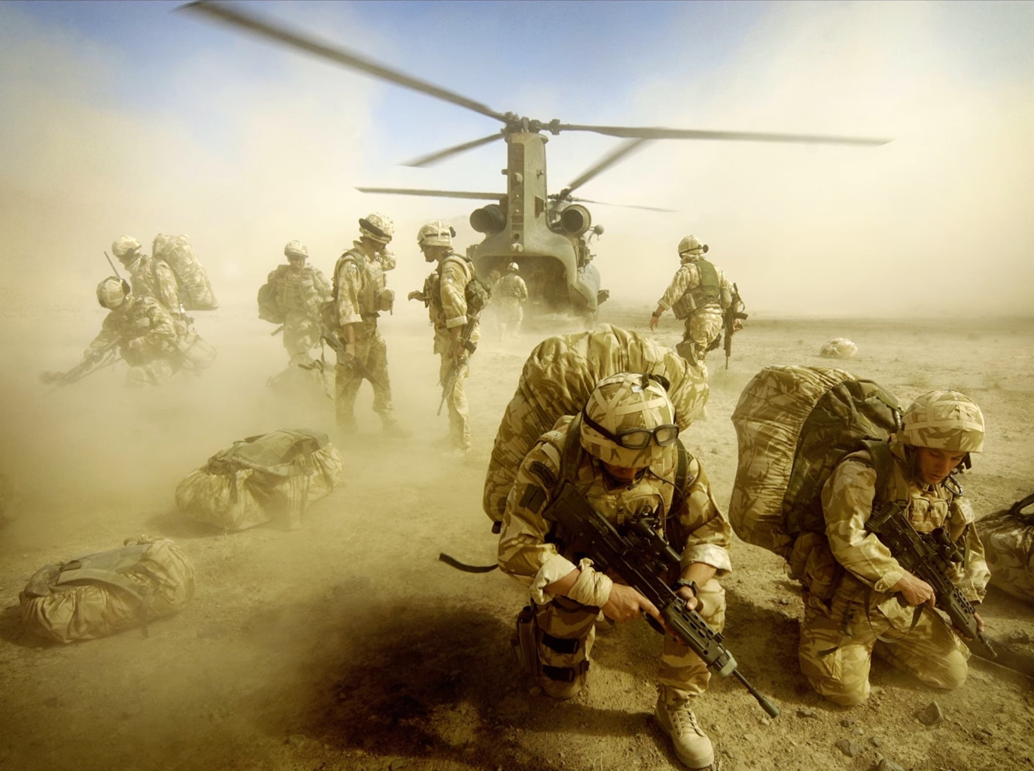 Soldiers in desert camouflage gear kneel and walk in a dusty landing zone near a parked Chinook helicopter.