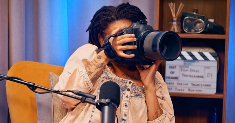 A person with dreadlocks holds a large camera up to their face, partially obscuring it, while sitting in front of a microphone and a film clapperboard on a shelf.