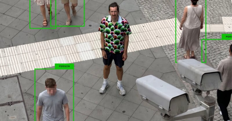 A high-angle shot shows several pedestrians walking on a paved city square, with a security camera in the foreground and bounding boxes overlaid on the people.