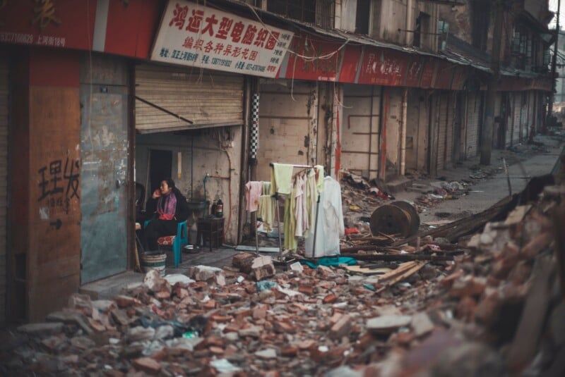A woman sits on a blue chair inside a partially demolished storefront, while a rack of laundry stands amidst rubble on the street.