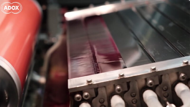 A close-up view of a machine coating a strip of dark, glossy film with a liquid substance, with a large red roller visible on the left.