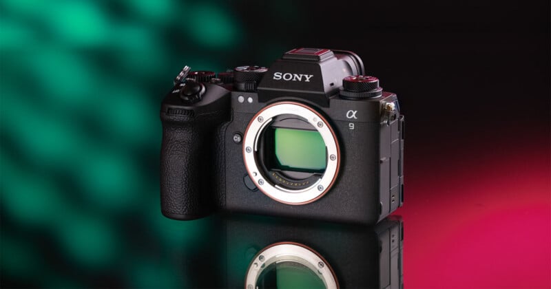 A black Sony Alpha 9 mirrorless camera body sits on a reflective surface against a background that transitions from green bokeh on the left to solid red on the right.