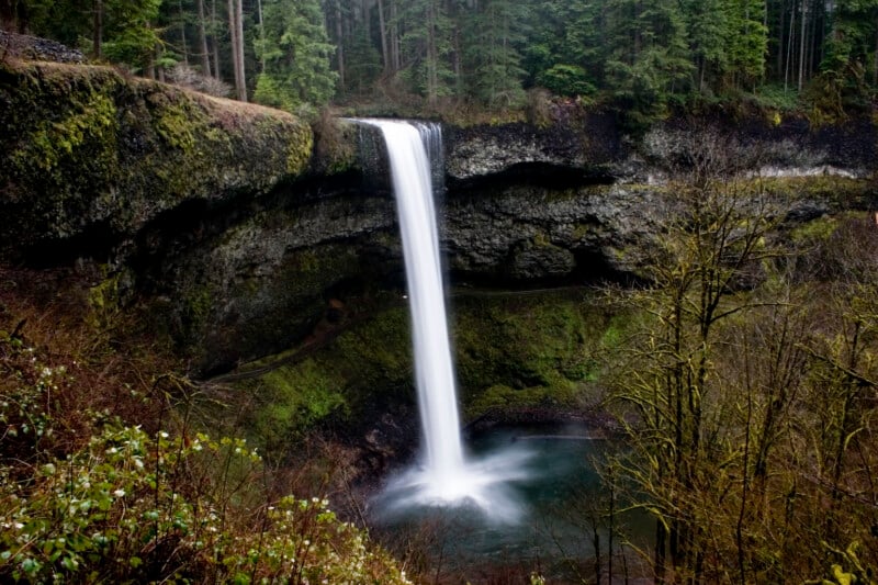 A tall, narrow waterfall plunges over a dark, mossy cliff into a pool surrounded by a dense forest.
