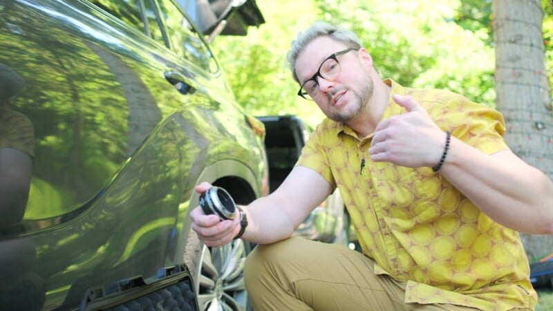 A man with blonde hair and glasses, wearing a yellow patterned shirt, kneels beside a car while holding a small black camera lens adapter.