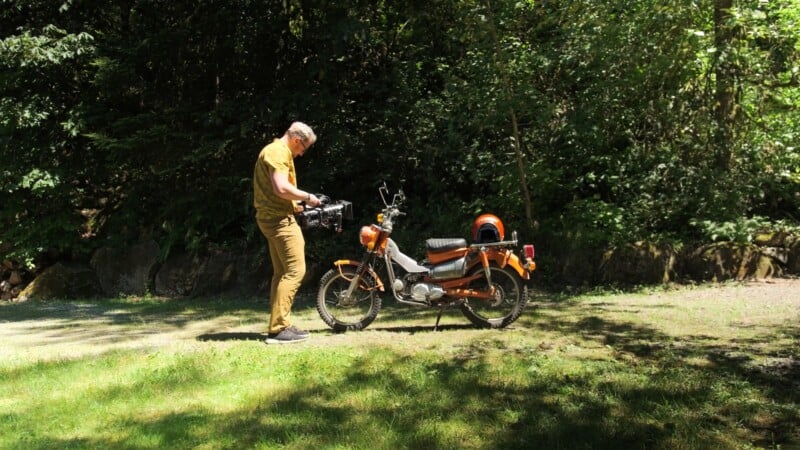A man in a yellow shirt and tan pants stands in a grassy clearing, holding a professional video camera while facing an orange vintage moped.
