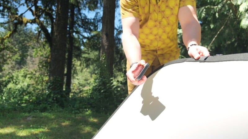 A person wearing a yellow patterned shirt holds a smartphone over a white reflector in a wooded area.