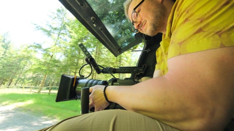 A man wearing glasses and a yellow patterned shirt holds a professional video camera while sitting in the back of a vehicle driving past trees.