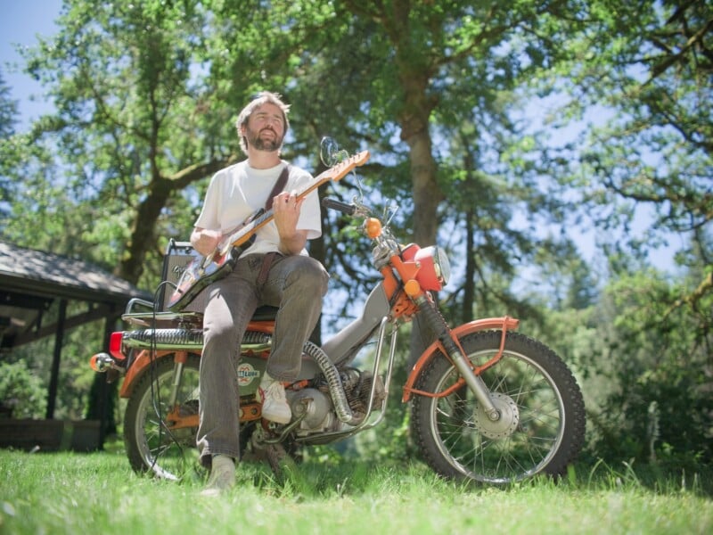 A man with a beard sits on an orange vintage motorcycle in a grassy field, playing an electric guitar connected to a small amplifier.