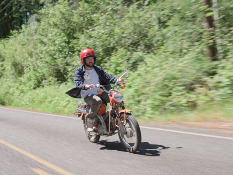 A man wearing a red helmet and dark jacket rides an orange vintage moped along a paved road lined with trees.