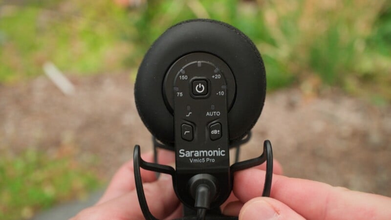 A close-up view of a Saramonic Vmic5 Pro microphone held in a person's hands, showing the rear control panel with power, gain, and filter settings.