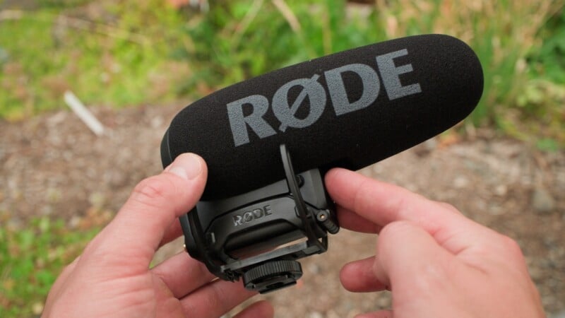 A person holds a black RØDE shotgun microphone with a foam windscreen in their hands.