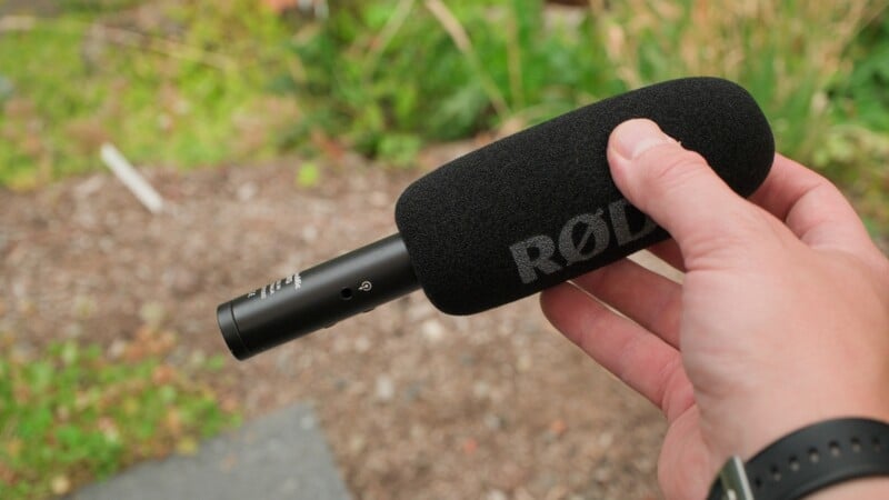 A hand holds a black RØDE shotgun microphone with a foam windscreen against a blurred outdoor background.