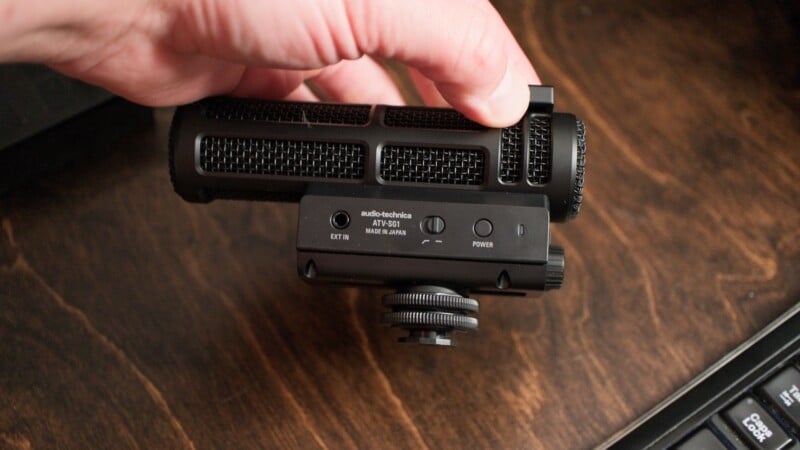 A hand holds an Audio-Technica ATV-SG1 shotgun microphone above a wooden desk surface next to a computer keyboard.