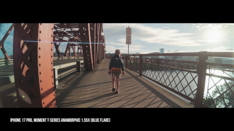 A child walks away from the camera along a pedestrian walkway on a red steel bridge, with a bright sun flare and city skyline in the distance.