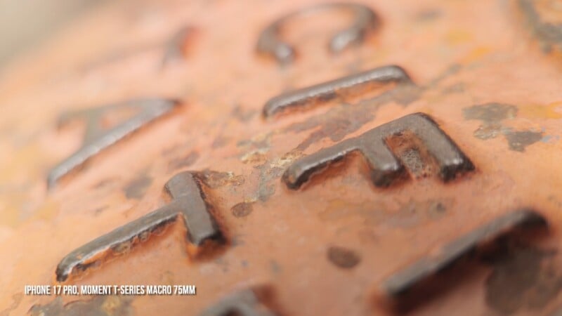 A close-up, macro view of weathered, orange metal with raised, dark, rusted lettering, including the visible text "IPHONE 17 PRO, MOMENT T-SERIES MACRO 75MM".