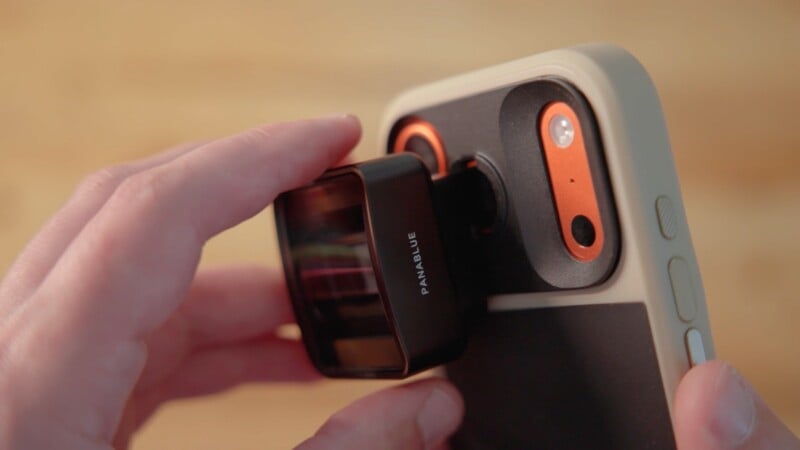 A hand attaches a black, rectangular "PANABLUE" branded lens accessory to the camera module of a smartphone in a light-colored case.