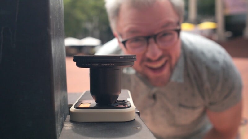 A man with glasses smiles behind a smartphone mounted with a large telephoto lens attachment.