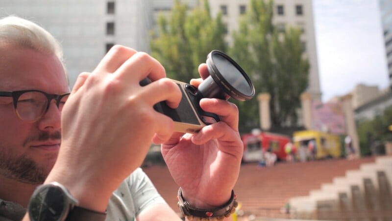 A person with blonde hair and glasses holds a smartphone equipped with a large circular lens filter in an outdoor urban setting.