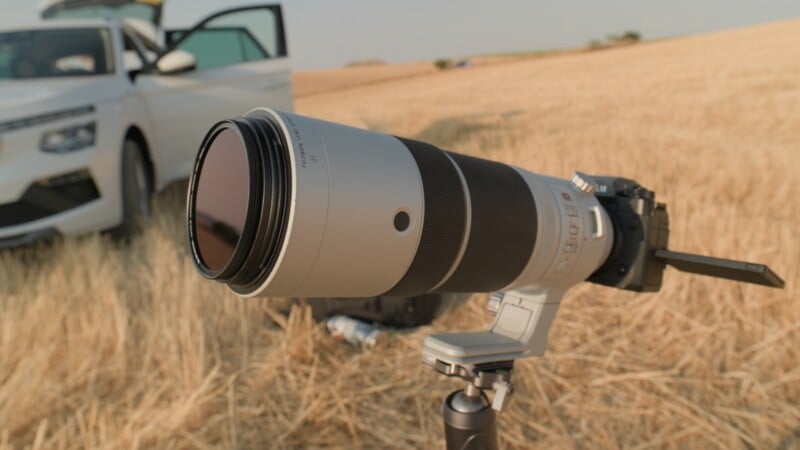 A long telephoto lens attached to a camera on a tripod stands in a dry, golden field with a white car parked in the background.