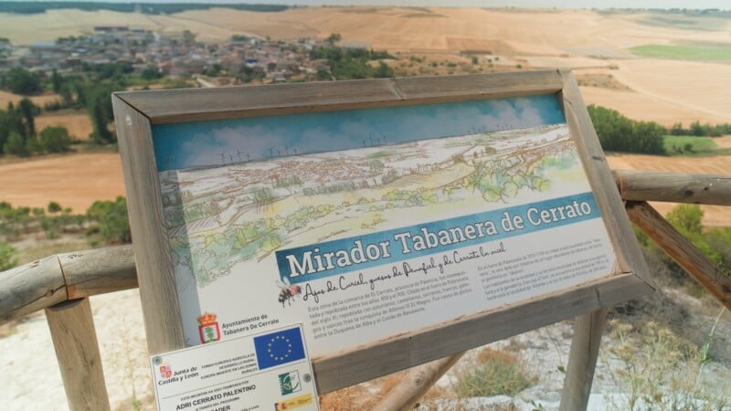 A wooden information sign stands on a hillside overlooking a small town and rural landscape, featuring a watercolor illustration and text about Mirador Tabanera de Cerrato.
