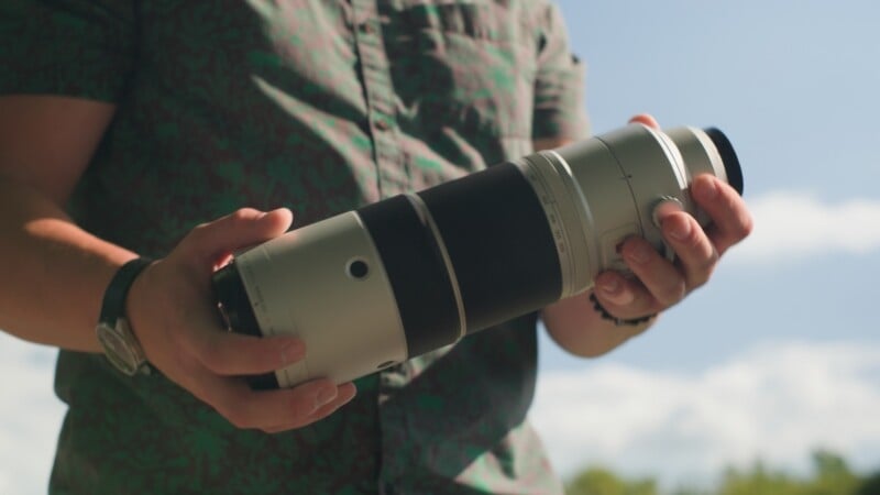 A person wearing a green patterned shirt holds a large, white telephoto camera lens with both hands.