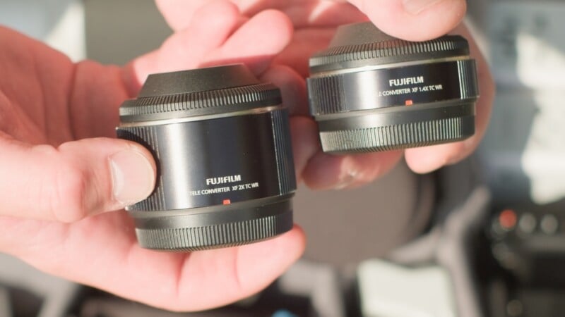 A person holds two Fujifilm teleconverters, a 2X TC WR on the left and a 1.4X TC WR on the right.