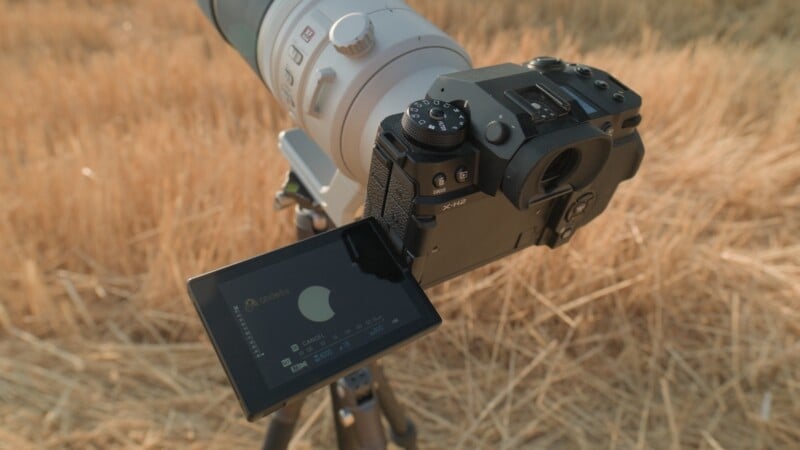 A Fujifilm X-H2 camera with a long telephoto lens is mounted on a tripod in a field of dry grass, displaying a partial solar eclipse on its screen.