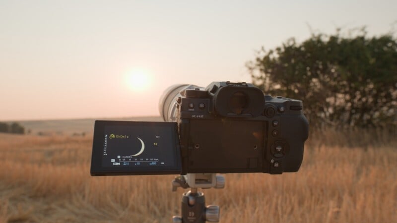 A Fujifilm X-H2 camera on a tripod in a field at sunset, with its screen displaying a crescent moon icon and exposure settings.