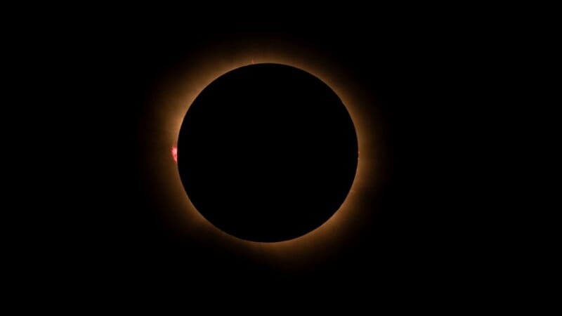 A total solar eclipse shows the moon silhouetted against the sun, with the solar corona and small red prominences visible around the edges.