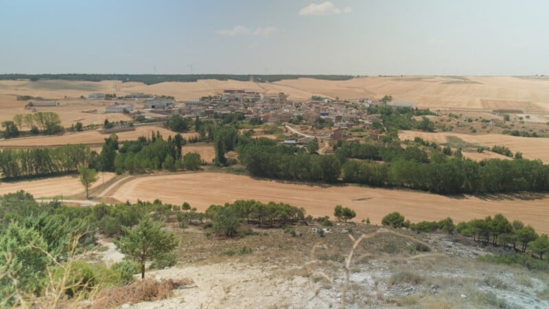 A small village sits nestled among golden agricultural fields and patches of green trees under a clear, hazy sky.