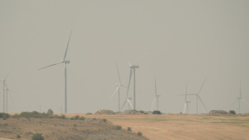 A row of wind turbines stands in a dry, golden field under a hazy, overcast sky.